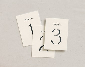 May include: Three ivory table number cards with black script. Each card has the word "table" written at the top, with the numbers 1, 2, and 3 in a bold, elegant font. The cards are arranged on a neutral surface.
