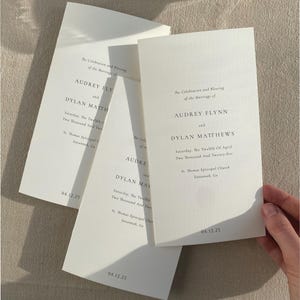 May include: Off-white wedding programs with black text. The programs are for Audrey Flynn and Dylan Matthews, with the date 04.12.25. The programs are stacked and fanned out, with one held by a hand.