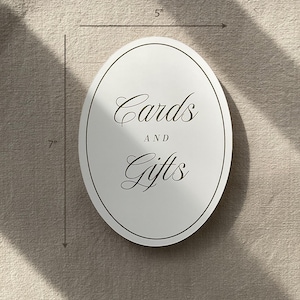 May include: An oval, white sign that is 7 inches tall and 5 inches wide, with the words "Cards AND Gifts" in elegant script. The sign has a thin, dark border.