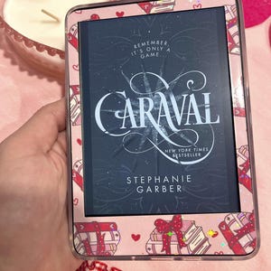 May include: A pink e-reader case with a repeating pattern of books and bows. The e-reader displays the book cover for "Caraval" by Stephanie Garber. The text "Remember, it's only a game..." is also visible.