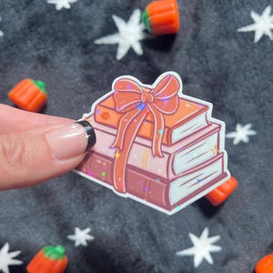 May include: A sticker featuring a stack of books with a large orange bow. The books are in shades of orange, pink, and brown. The sticker has a holographic effect. Scattered around are orange candy pumpkins on a dark gray background with white star shapes.