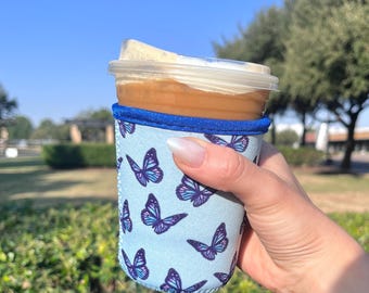 May include: A blue and purple butterfly patterned neoprene cup holder for a cold beverage. The cup holder is on a clear plastic cup with a brown beverage and white foam on top.
