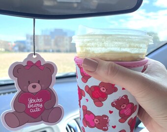 May include: A pink and white car air freshener with a cartoon bear holding a heart that says "You're Beary Cute". A pink and white cup cozy with a cartoon bear pattern is on a cup of coffee.