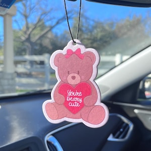 May include: A pink air freshener shaped like a teddy bear with a pink bow and a red heart. The heart has the text "You're beary cute" in white cursive. The air freshener hangs from a black string.