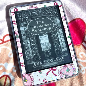 May include: An e-reader with a decorative cover featuring a Christmas theme. The screen displays the book cover of "The Christmas Bookshop" by Jenny Colgan. The cover design includes pink, white, and red accents with candy canes and Christmas tree illustrations.