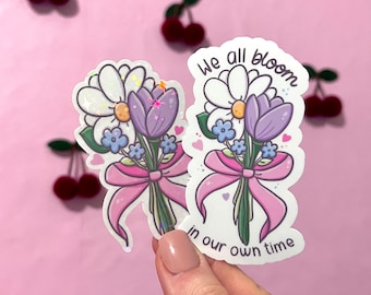 May include: Two white stickers with a bouquet of flowers, a daisy, a crocus, and small blue flowers tied with a pink ribbon. The text on the stickers reads "We all bloom in our own time".