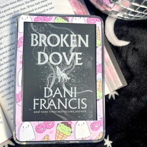 May include: An e-reader device featuring a protective case with a light pink background decorated with a pattern of cartoon ghosts, cherries, and ice cream cones. The screen displays the book cover for Broken Dove by Dani Francis.