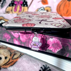 May include: A phone case with a pink and white pattern of pumpkins and bows, resting on a stack of books. A clear charm of a ghost wearing a scarf and holding a drink hangs from the case. The background is a pink Halloween-themed blanket.