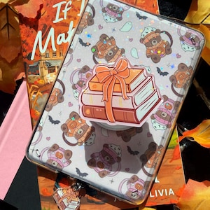 May include: A clear tablet case featuring a repeating pattern of brown teddy bears wearing pink and orange hats, small ghosts, and bats. A central sticker and a small hanging charm both depict a stack of three books tied with an orange ribbon.