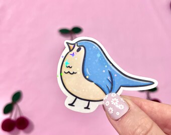 May include: A blue and white bird sticker with glitter accents. The bird is facing the right and has a small, round body with a long, pointed beak. The sticker is on a pink background.
