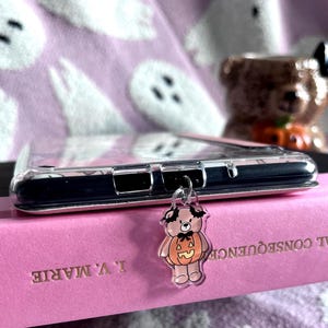 May include: A clear phone case featuring a small, dangling acrylic charm shaped like a light brown teddy bear wearing a pumpkin costume with bat wings. The charm is attached to the bottom of the phone case with a silver metal jump ring.
