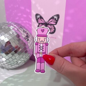 May include: A pink sticker of a cartoon nutcracker soldier with a heart on his hat. The nutcracker has a white mustache and is wearing a pink uniform. A disco ball and a pink butterfly card are in the background.