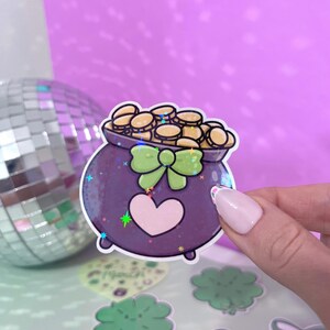 May include: A purple cauldron filled with gold coins, decorated with a green bow and a pink heart. The cauldron is a sticker with a glittery finish.
