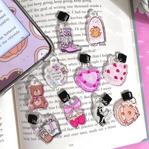 May include: A set of six clear plastic bookmark charms with various cute illustrations. The charms feature a ghost holding a pumpkin, a pair of pink cowboy boots, a teddy bear, a heart with strawberries, a book with a coffee cup, and a strawberry with a bow. Each charm has a black clip to attach to a book.