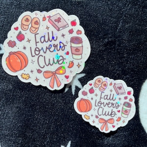 May include: Two stickers with a fall theme. The larger sticker features a pumpkin, coffee cup, book, and bow, with the text "Fall Lovers Club." The smaller sticker has the same design. Both stickers have a holographic finish.