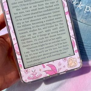 May include: An e-reader device featuring a clear protective case decorated with a pattern of pink hearts, moths, and dragon wings. The screen displays black text on a white background, showing a narrative passage and 12 mins left in chapter.
