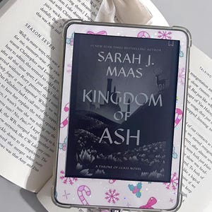 May include: An e-reader with a decorative cover featuring a pattern of pink candy canes, snowflakes, and bows. The screen displays the book "Kingdom of Ash" by Sarah J. Maas. A pink charm hangs from the bottom.