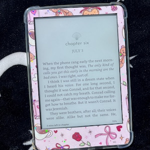 May include: A digital e-reader with a pink cover featuring teddy bears, bows, and roses. The screen displays text from a book, with the chapter title "chapter six" and the date "JULY 3" visible. The e-reader is on a black and white star patterned surface.