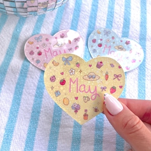 May include: Three heart-shaped stickers with glitter and various illustrations. The stickers are yellow, pink, and blue. Each sticker has the word "May" written on it.