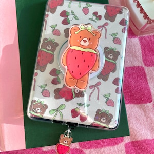 May include: Clear phone case with a repeating pattern of brown bears wearing strawberry costumes and green bows. A large bear with a strawberry costume and a bow is attached to the case. A small bear charm with a strawberry costume hangs from the bottom.