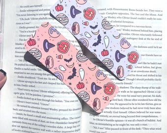 May include: Two Halloween-themed bookmarks with a repeating pattern of pumpkins, witch hats, bats, and ghosts in pink and lavender. The bookmarks are placed on top of an open book.