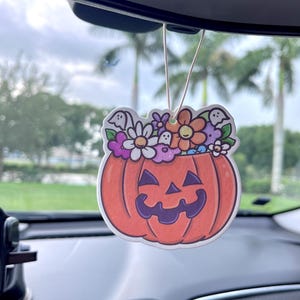 May include: A pumpkin-shaped car air freshener with a smiling jack-o'-lantern face. The pumpkin is orange with a purple mouth and is topped with colorful flowers and ghost figures. The air freshener hangs from a string.