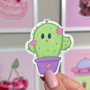 Op de afbeelding: Een groene cactus met een glimlachend gezicht en roze bloemen in een paarse pot met een hartje op de voorkant. De cactus heeft glitter.