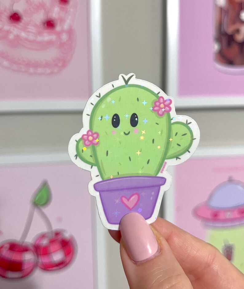 Op de afbeelding: Een groene cactus met een glimlachend gezicht en roze bloemen in een paarse pot met een hartje op de voorkant. De cactus heeft glitter.