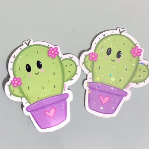 Op de afbeelding: Twee groene cactus stickers met roze bloemen en paarse potten. De cactussen hebben lachende gezichten en zijn in cartoon stijl.