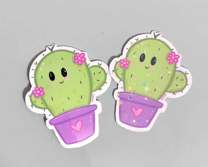 Op de afbeelding: Twee groene cactus stickers met roze bloemen en paarse potten. De cactussen hebben lachende gezichten en zijn in cartoon stijl.