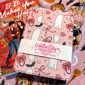 May include: A pink Kindle case with a Halloween-themed pattern featuring ghosts, bats, pumpkins, and bows. The case is labeled "Kindle Case" and "Kindle Basic 2022 & 2024". A book titled "If It Makes You Happy" is also visible.