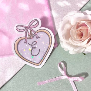 May include: A heart-shaped sticker with a gold frame and a pink bow. The heart is filled with a glittery purple background and the letter "E" is written in white inside the heart.