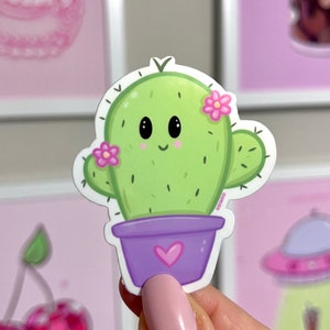 Op de afbeelding: Een groene cactus met een glimlachend gezicht en roze bloemen in een paarse pot met een roze hart. De cactus is een sticker.