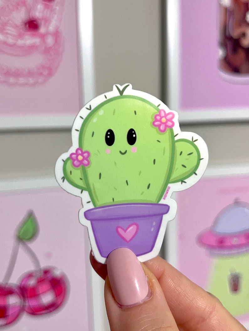 Op de afbeelding: Een groene cactus met een glimlachend gezicht en roze bloemen in een paarse pot met een roze hart. De cactus is een sticker.