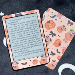 May include: An e-reader device and a matching protective cover featuring a repeating pattern of brown teddy bears wearing orange jack-o'-lantern costumes, black bows, and bats on a light pink background with a subtle holographic glitter effect.