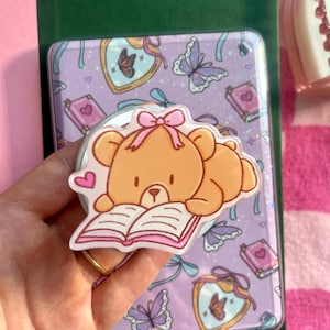 May include: A light brown bear-shaped phone grip with a pink bow, reading a book. The grip is on a lavender phone case with butterfly and book designs. The case is on a green surface.