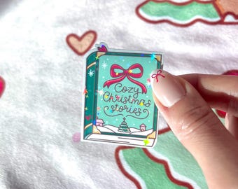 May include: A sticker in the shape of a book with the words "Cozy Christmas Stories" on the cover. The book is teal with a red bow and a winter scene. The sticker has a holographic effect.