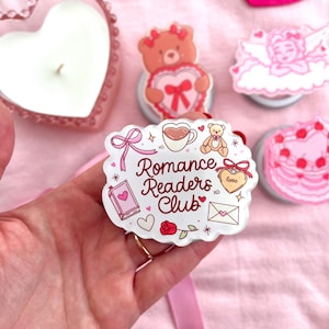 May include: A hand holds a white and pink Romance Readers Club sticker with illustrations of a book, rose, teddy bear, and coffee cup. Other pink heart-shaped items are in the background, including a candle and pop sockets.