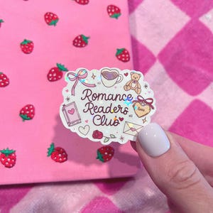 Peut inclure: Un autocollant avec le texte "Romance Readers Club" et diverses illustrations, dont un cœur, un livre, un ours en peluche et une tasse à café. L'autocollant est posé sur un fond rose avec des motifs de fraises et de cœurs.