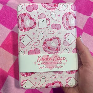 May include: A pink Kindle case featuring a pattern of pink cherries, heart-shaped cakes, ghost figures, and mugs. The case has a white band with the text "Kindle Case" and "Just one more chapter."