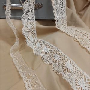 May include: Three white lace trims with different patterns. The lace trims are on a beige fabric background.