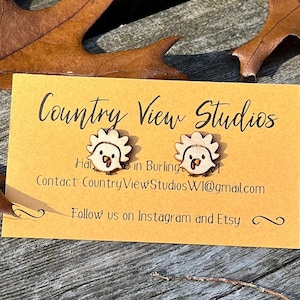 May include: Wooden turkey earrings with a cartoon design. The earrings are on a yellow card with the text "Country View Studios". The background includes brown leaves and a wooden surface.