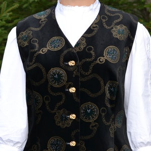 May include: Black velvet vest with a gold and teal clock and chain pattern. The vest has gold buttons and a V-neck. The vest is worn over a white blouse with a ruffled collar and lace sleeves. The vest is part of a vintage clothing collection.