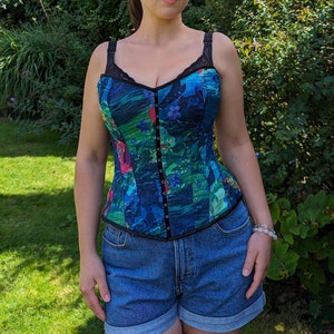 May include: A colorful corset top with a floral pattern in shades of blue, green, and pink. The top has black trim, straps, and a lace neckline. It features a hook-and-eye closure down the front.