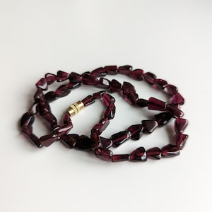 Peut inclure: Un collier composé de perles rouge foncé de forme irrégulière. Les perles sont enfilées, formant un collier avec un fermoir doré. Le collier est présenté sur une surface blanche.