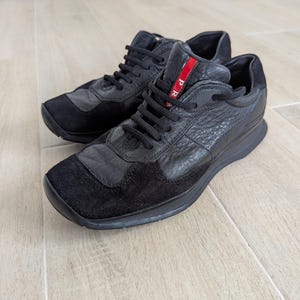 Puede incluir: Zapatillas deportivas negras con una parte superior de cuero texturizado y detalles de ante. Los zapatos tienen cordones negros y una franja roja con texto blanco. Las suelas son negras.