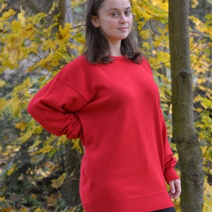 May include: A woman wearing a red sweater and black pants stands in a forest with yellow leaves.