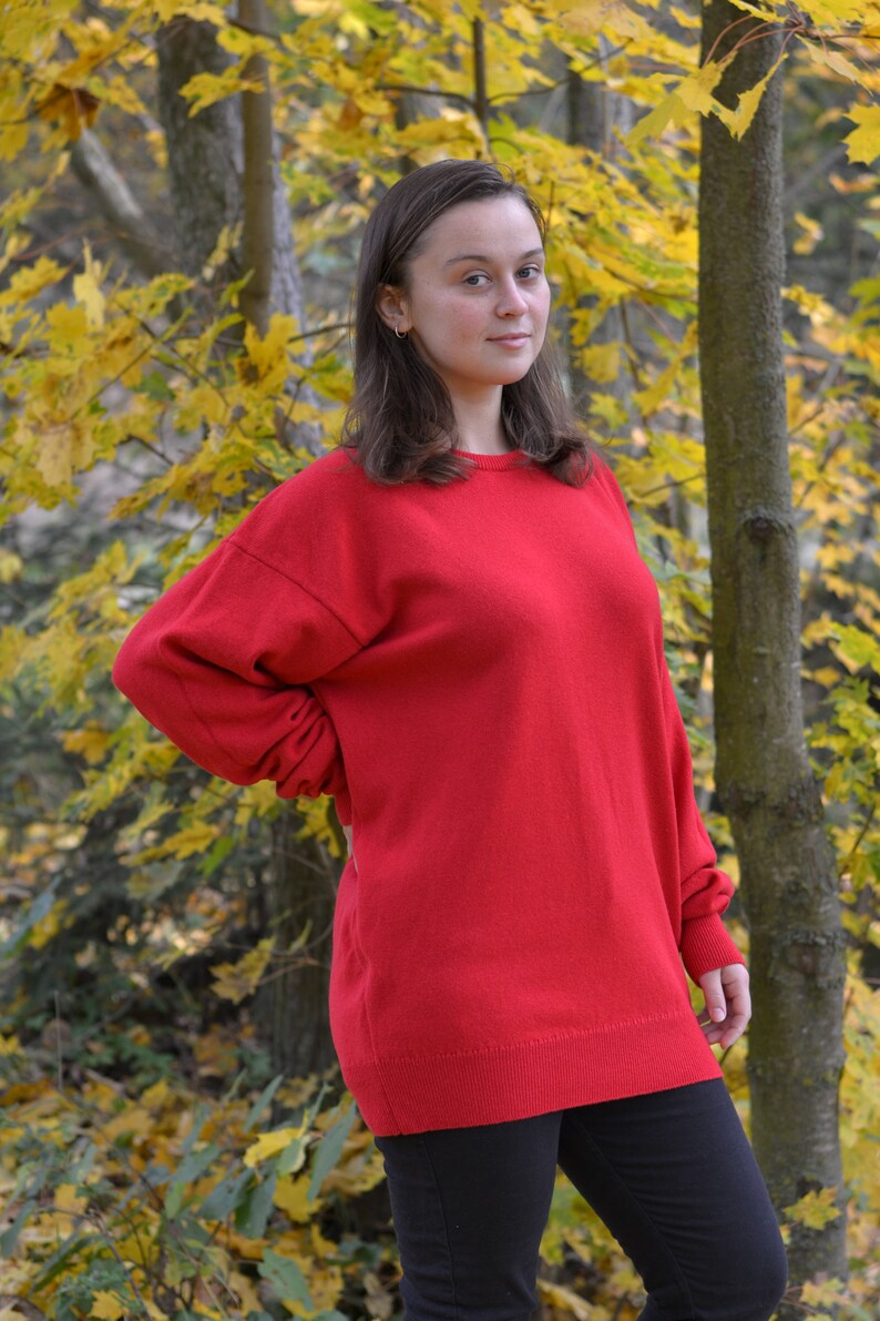 May include: A woman wearing a red sweater and black pants stands in a forest with yellow leaves.
