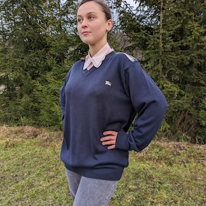 May include: A navy blue V-neck sweater with a small logo on the chest. The sweater is worn over a pink collared shirt and paired with gray jeans. The model is standing outside in front of green trees.