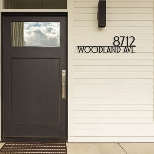 May include: A black metal house number sign with the number 8712 and the street name "WOODLAND AVE" on a white house exterior.